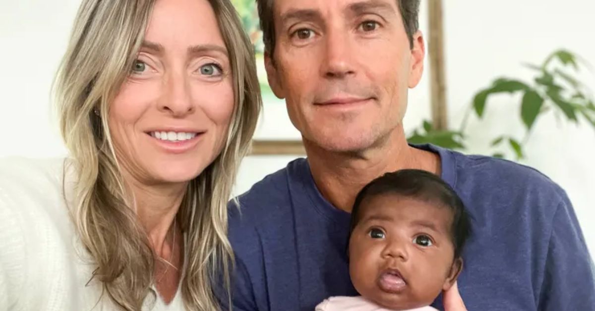 Family portrait featuring a smiling couple with their newborn baby, set against a bright, plant-filled background.