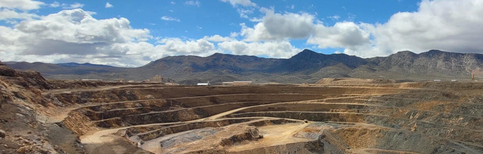 Expansive view of a mining site with layered earth formations and mountains under a partly cloudy sky.