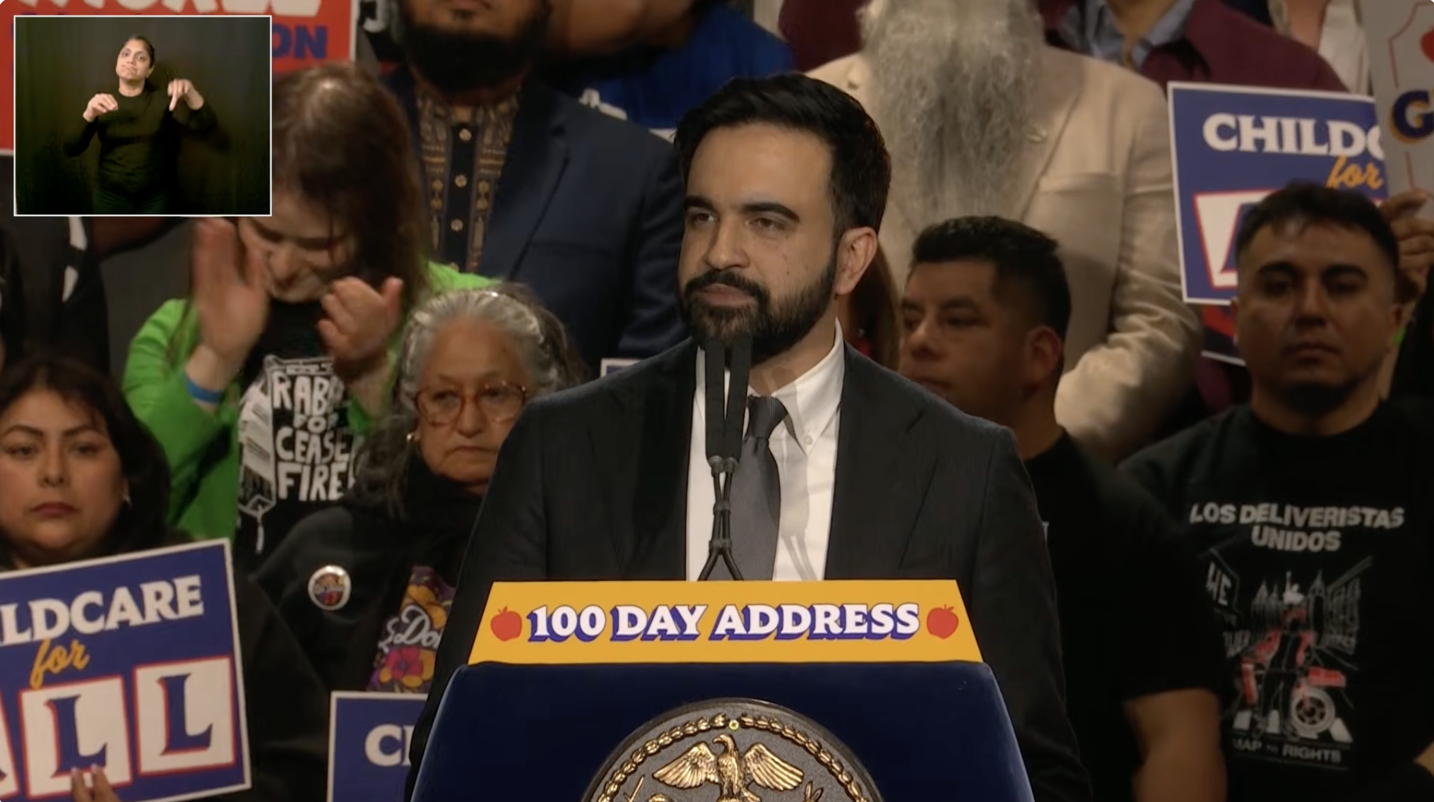 Speaker addressing the audience at a community event, with supporters holding signs advocating for childcare and social issues in the background.