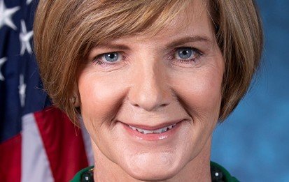 Professional headshot of a woman with short hair, smiling against a blue background, with an American flag visible in the backdrop.