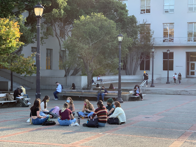 Students gather in a campus courtyard, sitting on the ground and engaging in discussion, with some individuals studying in the background.