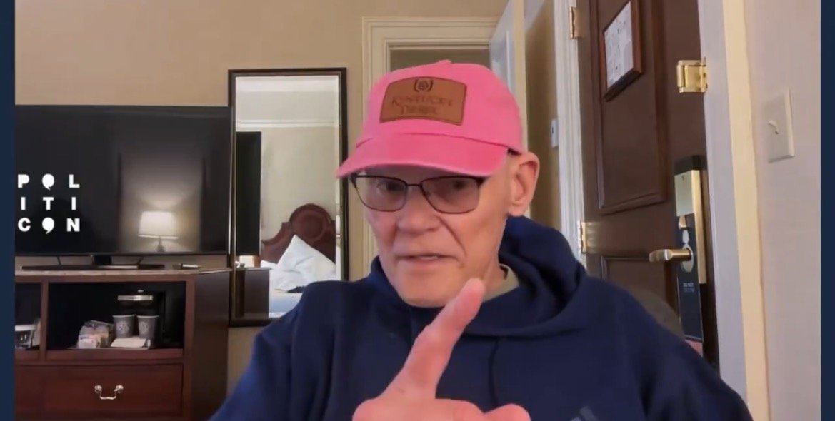 Person wearing a pink cap and glasses gestures while discussing topics at Politicon, with a hotel room and television in the background.
