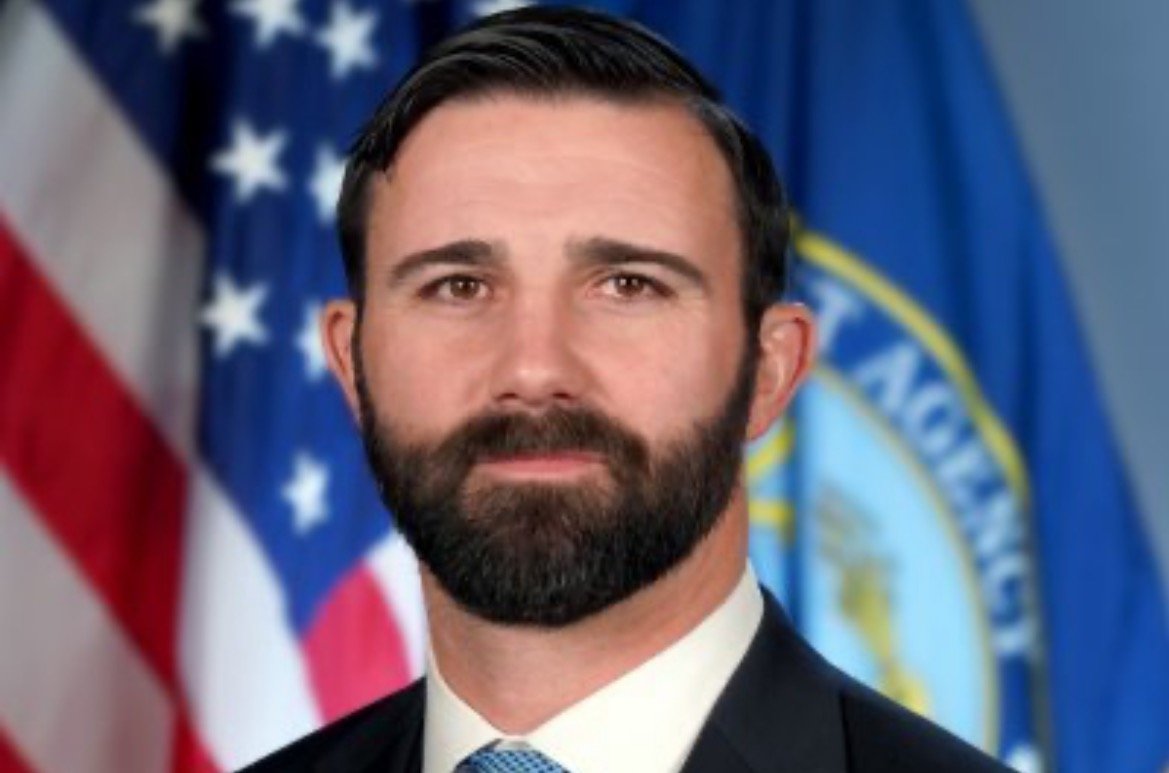 Professional portrait of a man with a beard in front of American and agency flags, representing leadership and authority.