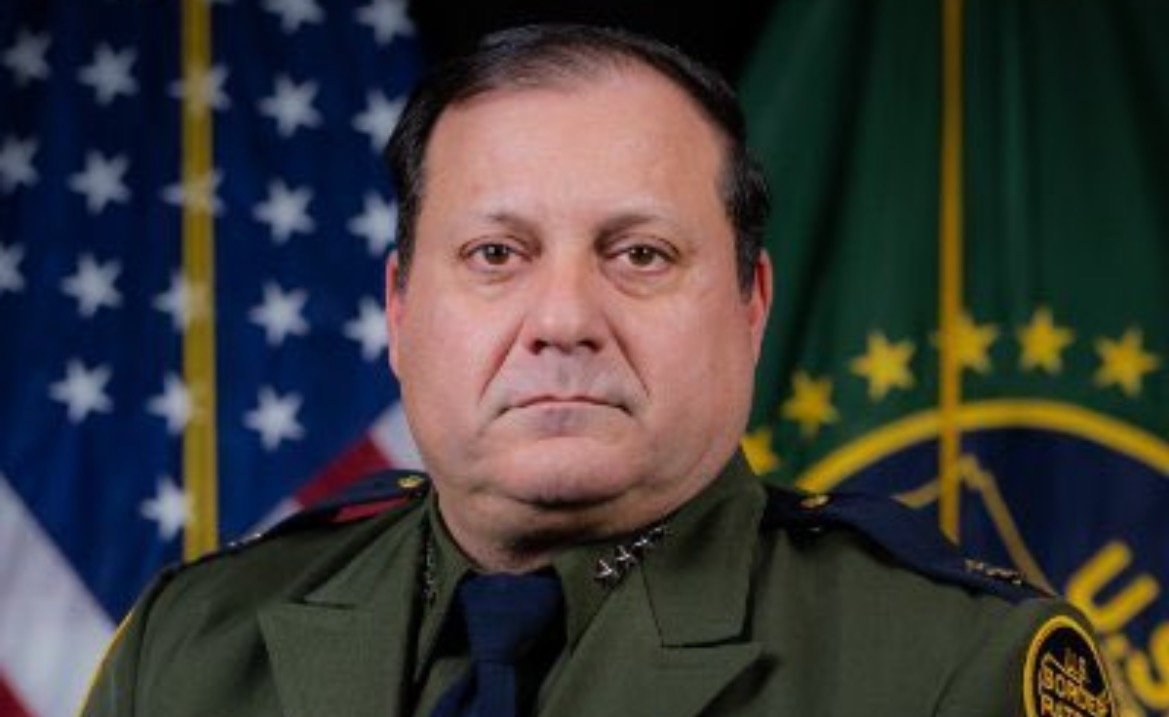 Portrait of a law enforcement officer in uniform, standing in front of the American flag and a departmental emblem, conveying authority and professionalism.
