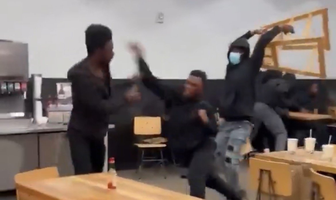 A chaotic scene in a cafeteria with individuals in black clothing engaging in a physical altercation, chairs overturned and drinks scattered.