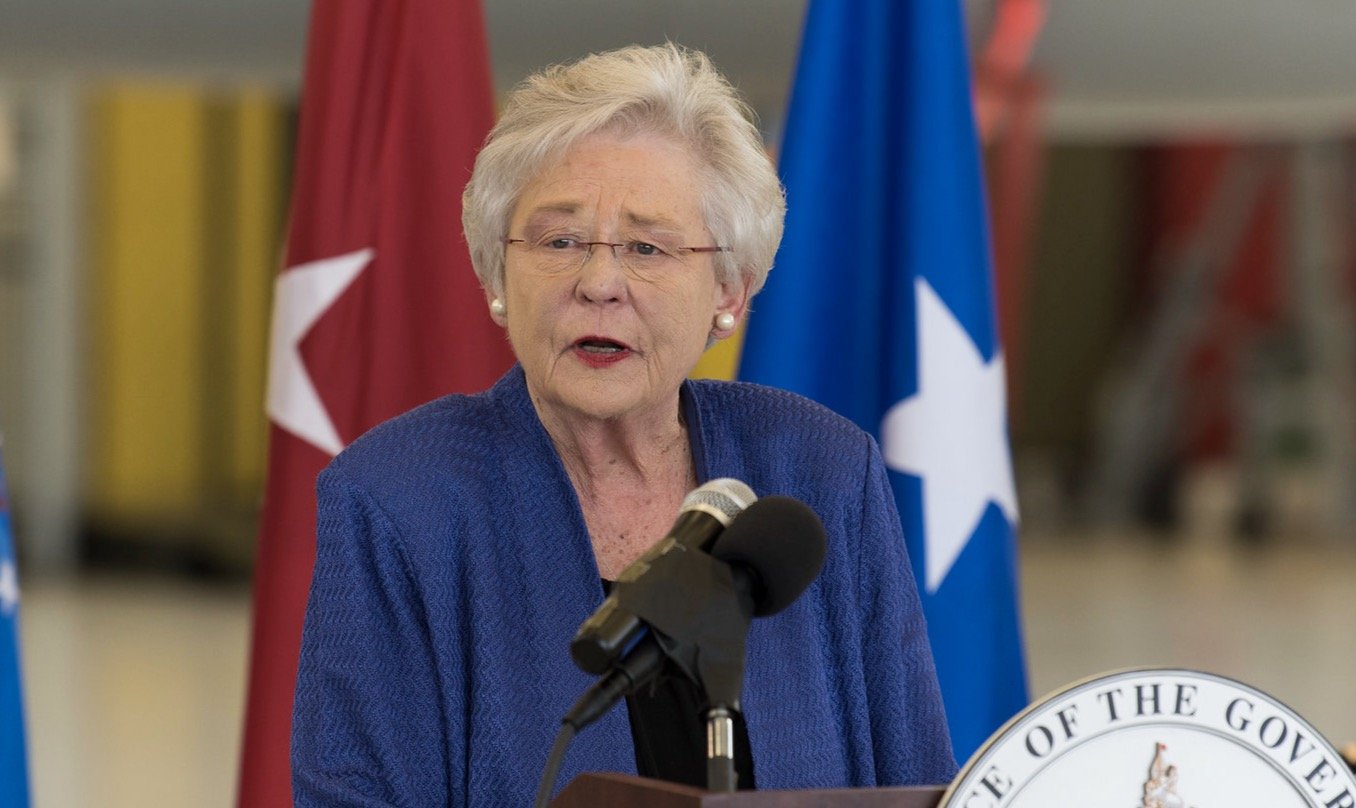 A woman in a blue jacket speaks at a podium with state seals and flags in the background, addressing an audience at an official event.