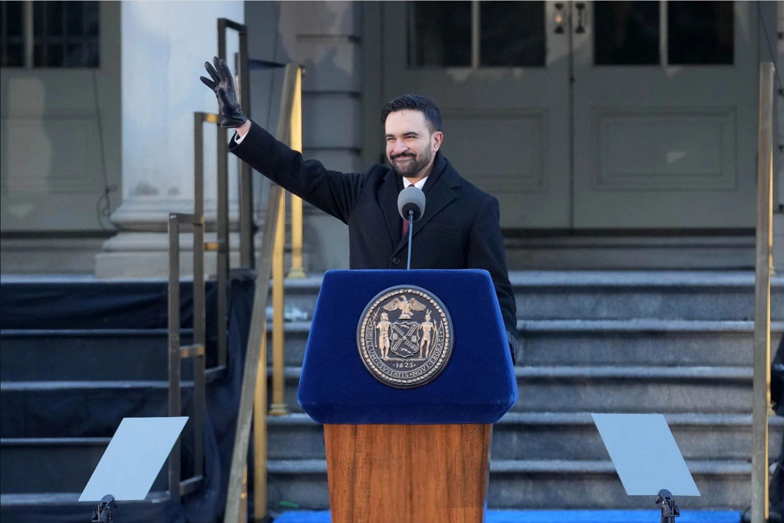 Man in a black coat and gloves waves from a podium with a city seal during an outdoor event.