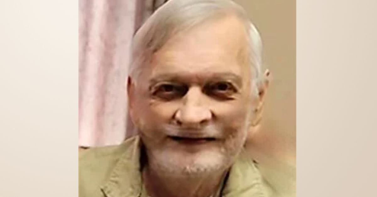 Smiling elderly man with gray hair and a beard, wearing a light-colored jacket, seated in a cozy indoor setting.