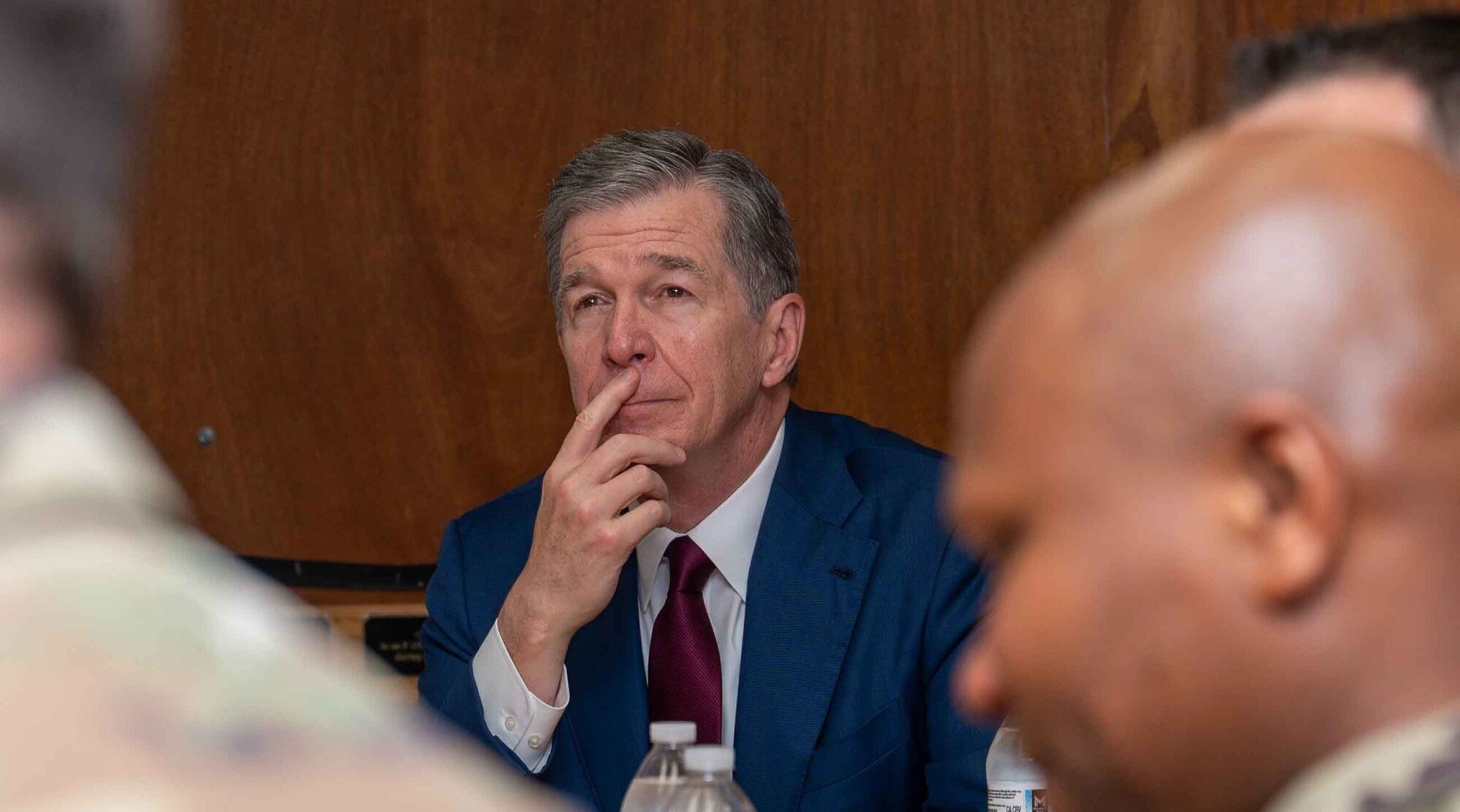 Man in a blue suit with a maroon tie thoughtfully listening during a meeting, surrounded by blurred figures in military attire.