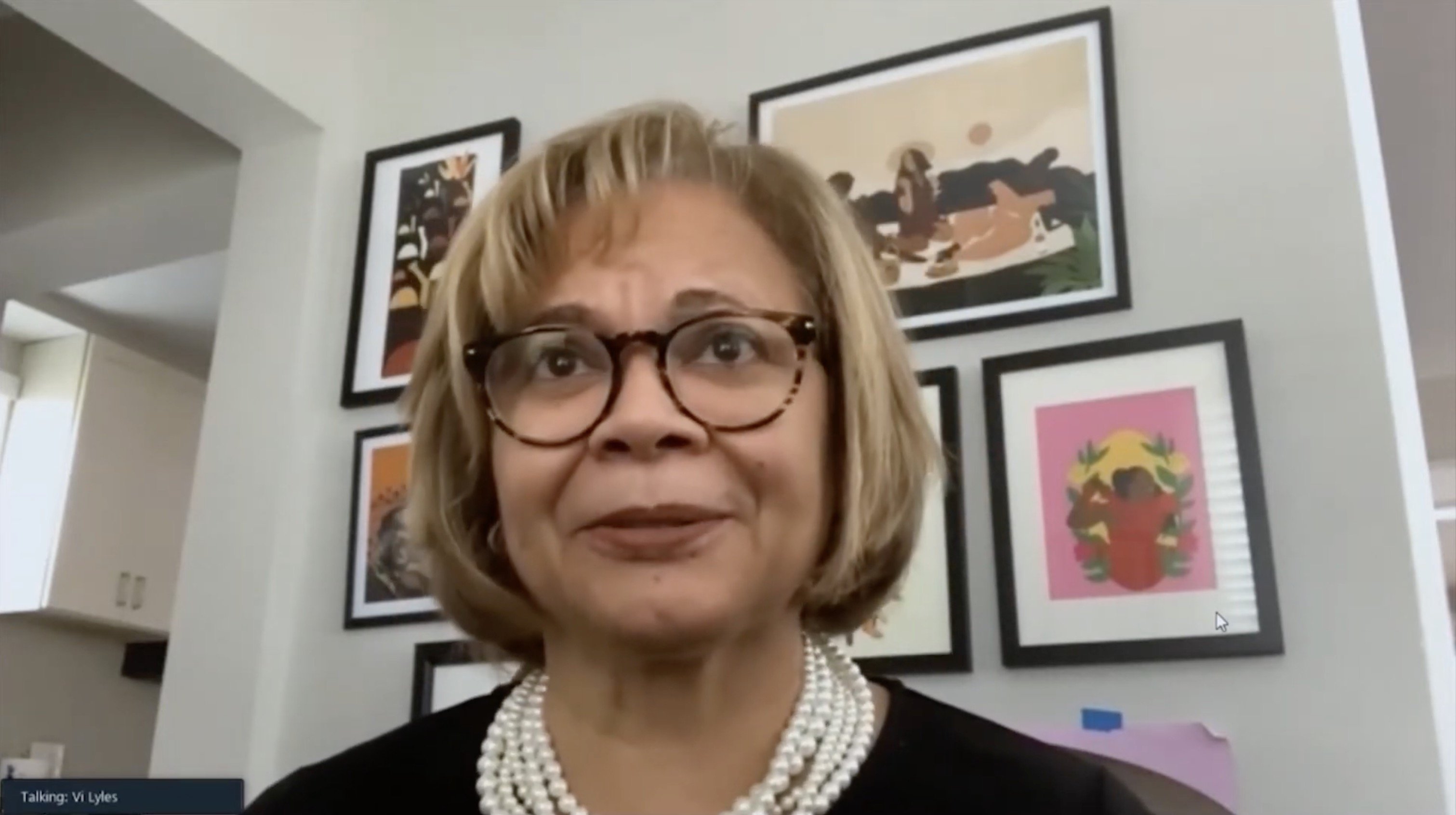 A woman with glasses and a pearl necklace smiles while seated in front of colorful artwork and a modern interior setting.