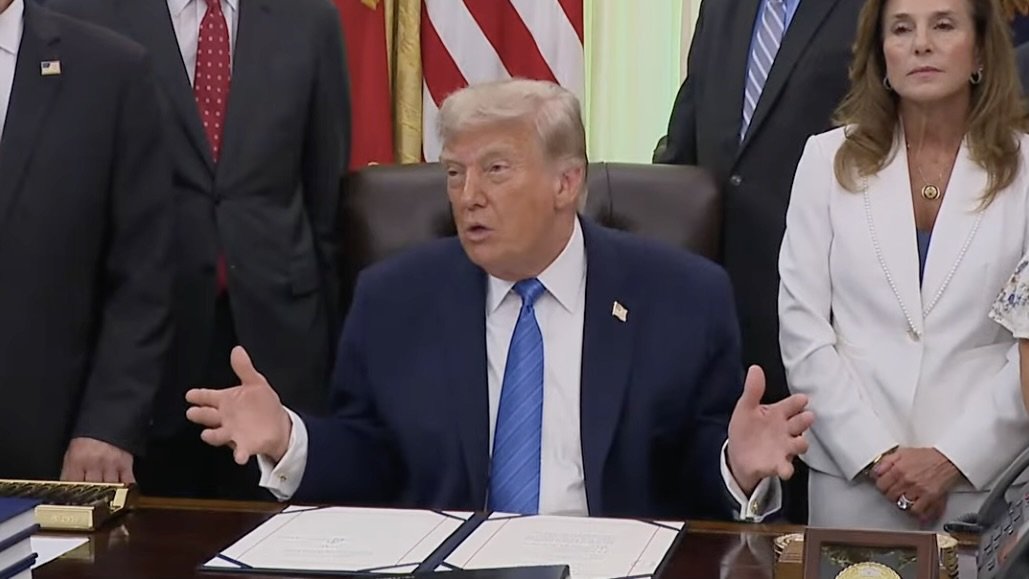 Donald Trump gestures while speaking at a desk surrounded by officials, with American flags in the background during a formal event.