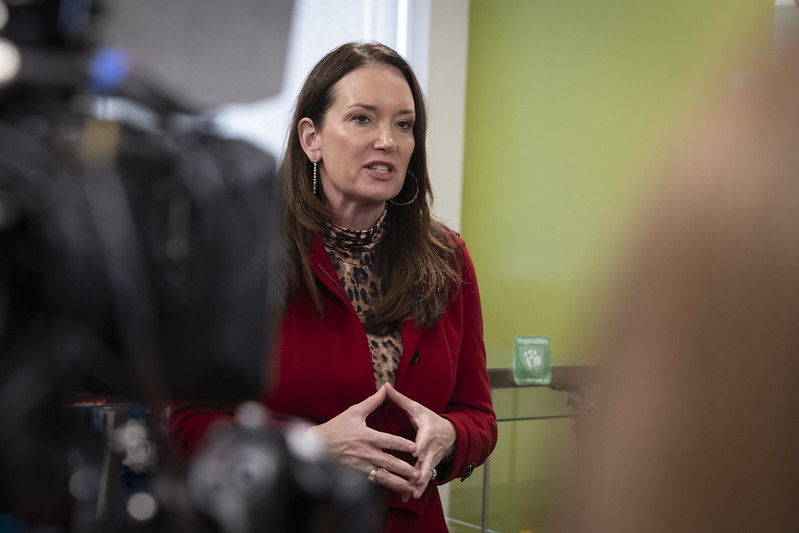 Woman in a red jacket speaks during a press event, with cameras capturing her presentation in a modern setting.