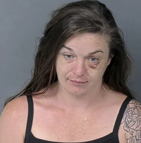 Booking mugshot of a woman with dark hair, blue eyes, and a bruise under her left eye, a tattoo visible on her shoulder.
