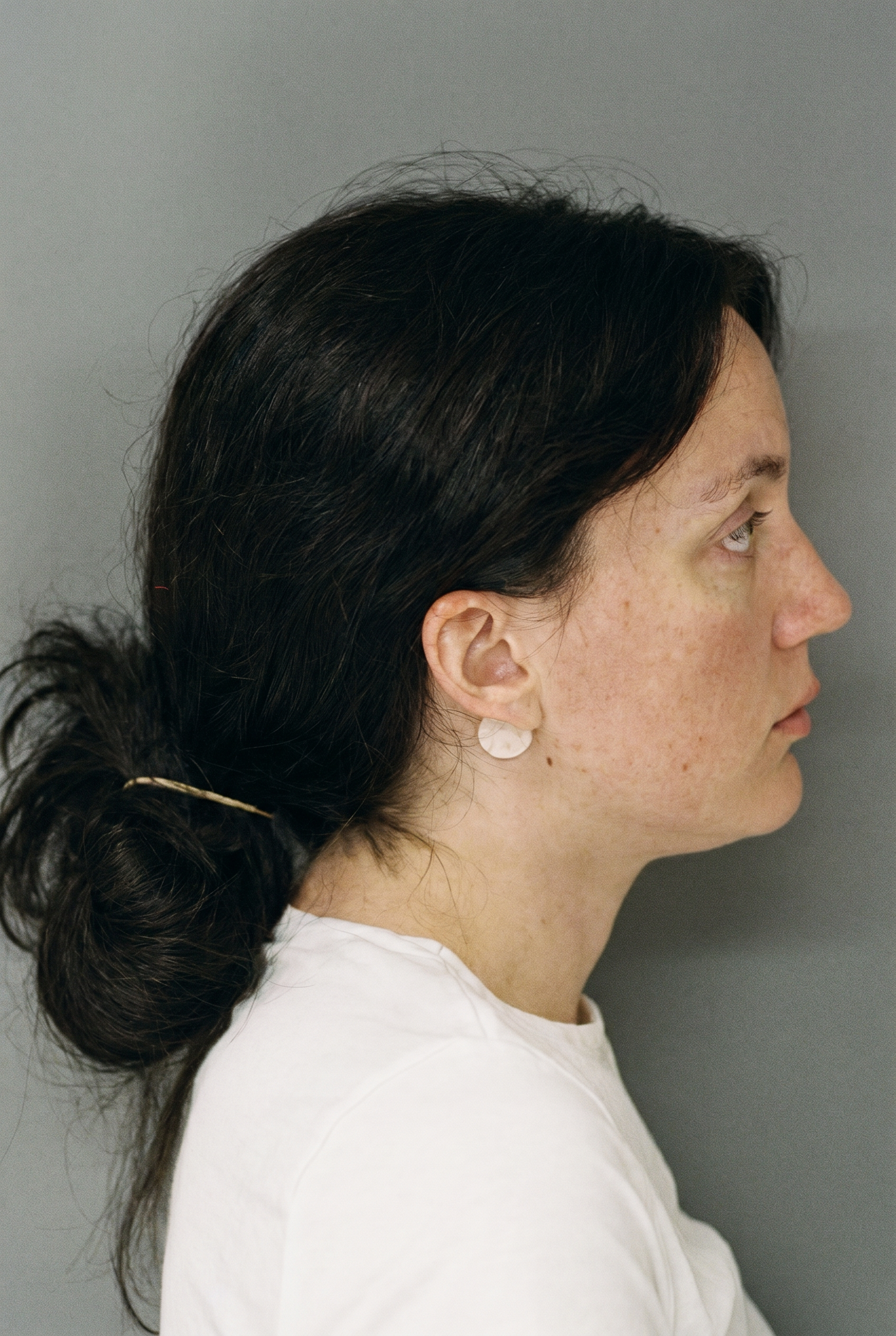 Side profile of a dark-haired woman in a white top wearing a circular earring, against a gray background.
