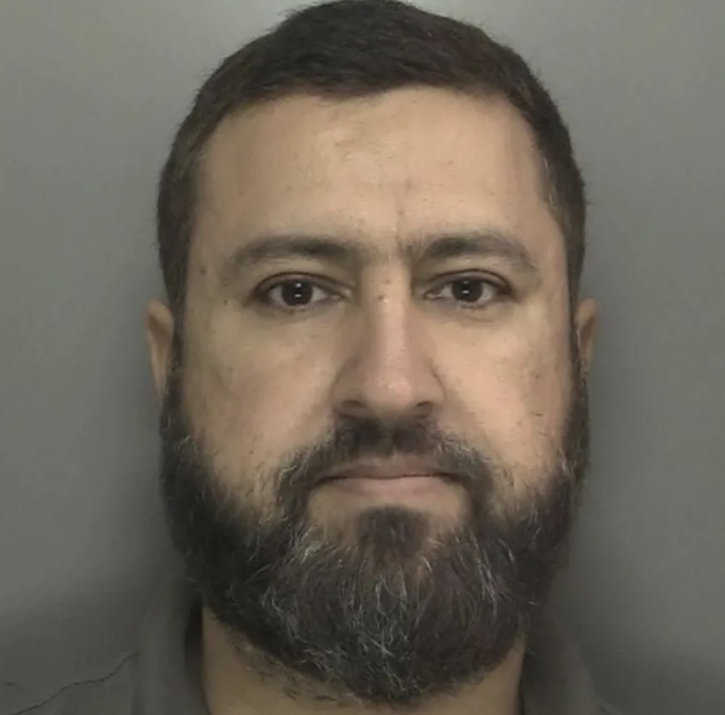 Police mugshot of a dark-haired man with a beard against a gray background.