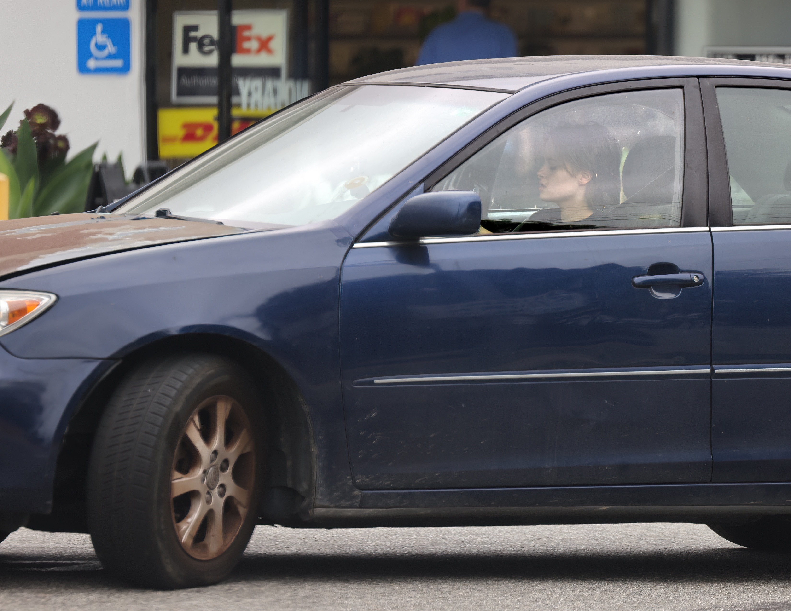 A young person visible through the passenger-side window of a beat-up navy blue Toyota sedan parked near a strip mall with FedEx and DHL signage.