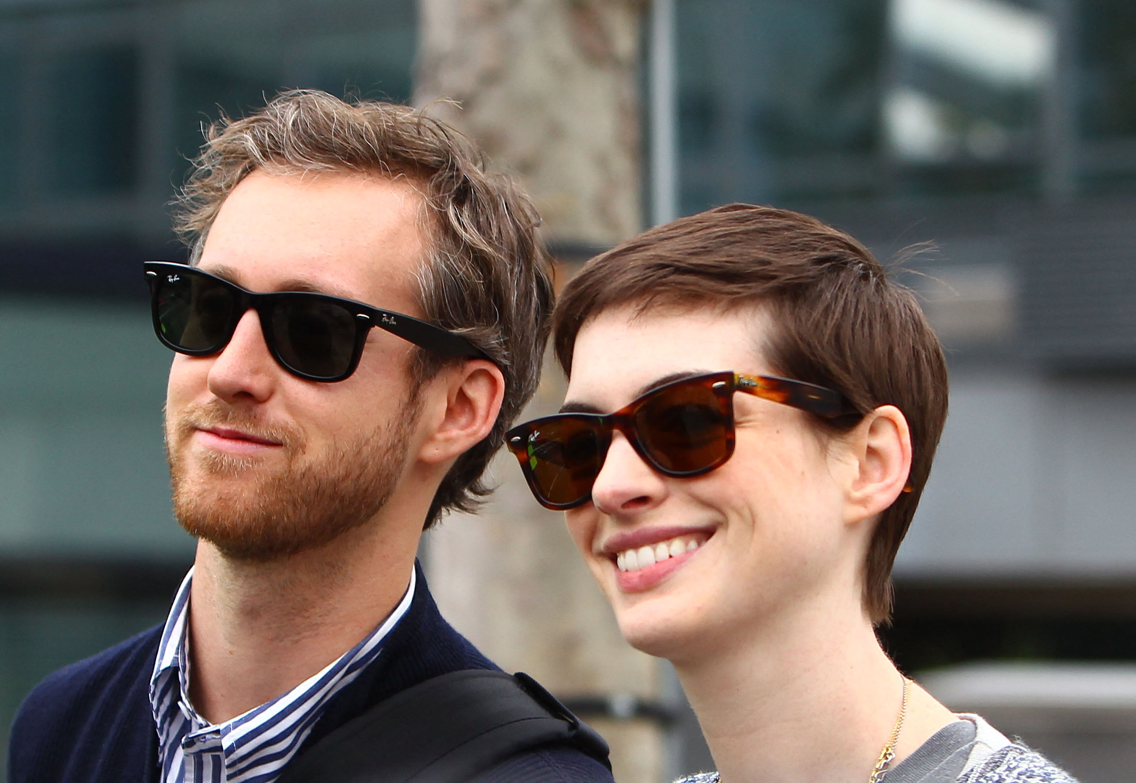 A man and short-haired woman wearing matching Ray-Ban sunglasses pose together outdoors, smiling.