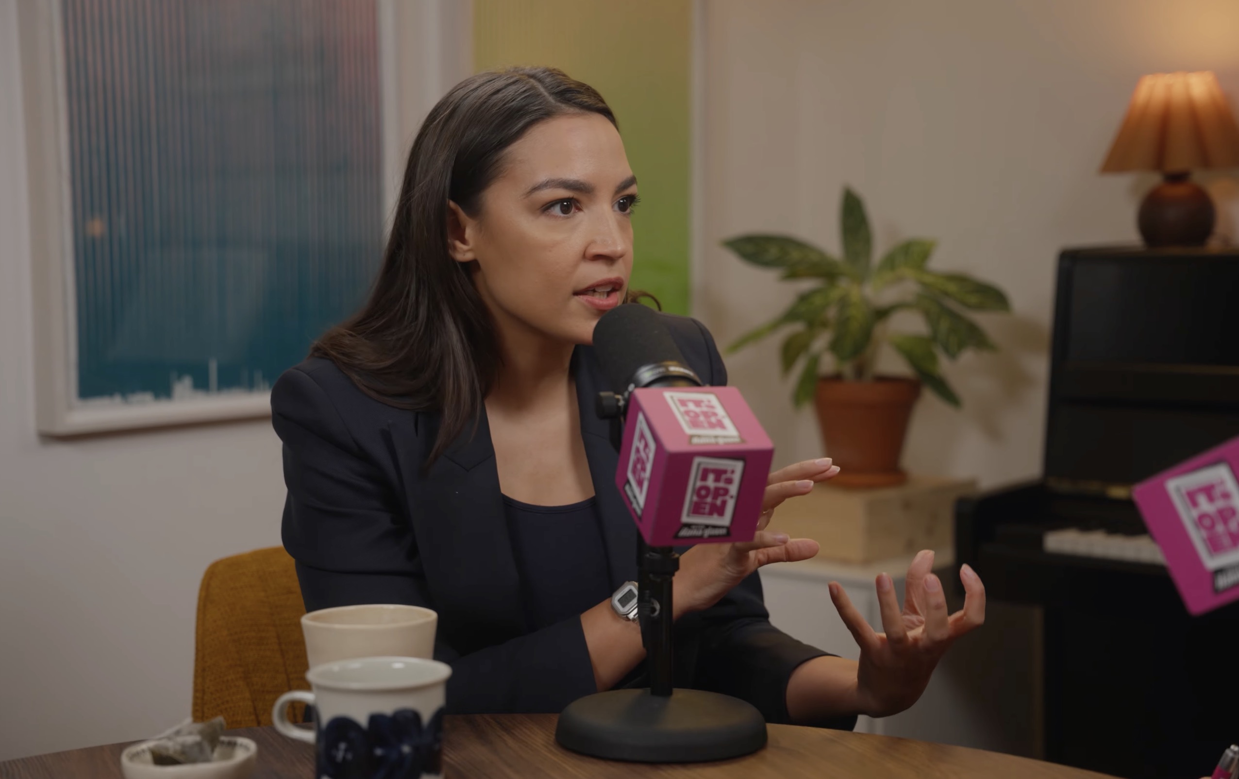 Woman in a dark blazer speaking into a pink cube microphone labeled 'It's Open' at a podcast studio table with coffee cups.