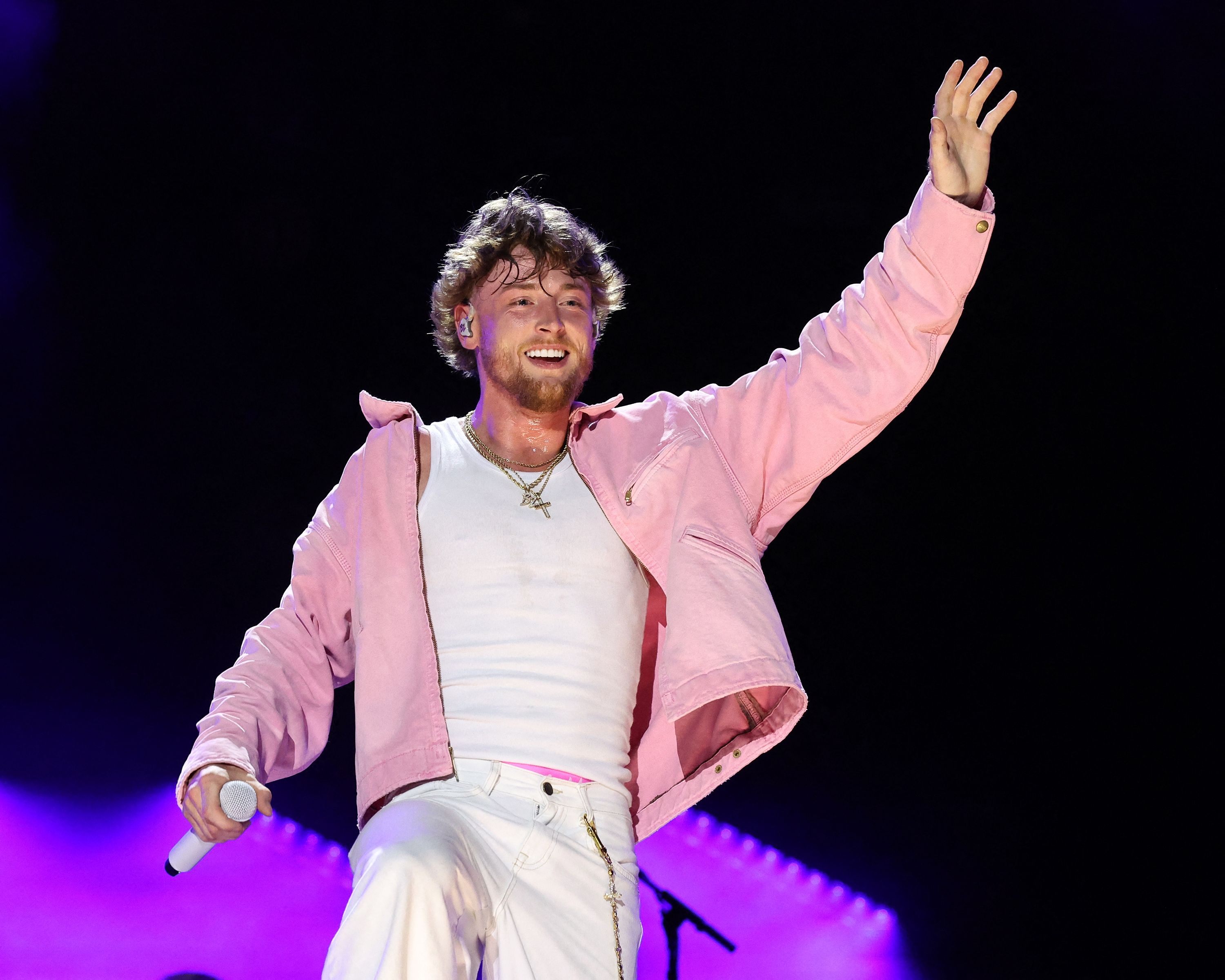 Male performer in a pink jacket and white tank top smiling and waving one arm in the air while holding a microphone on a darkened stage.