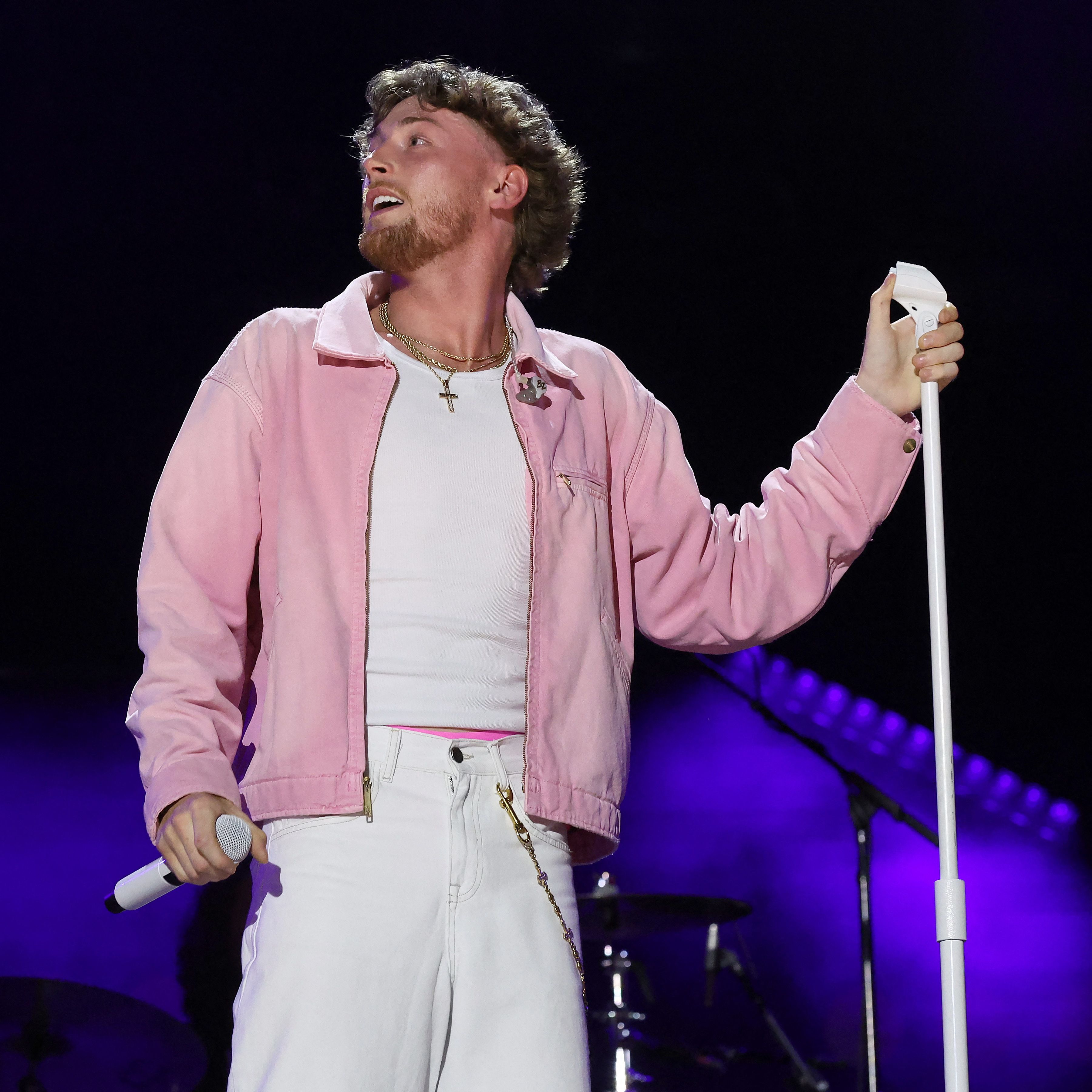 Male performer in a pink jacket and white jeans looking upward while holding a microphone in one hand and a mic stand in the other on a darkened stage.