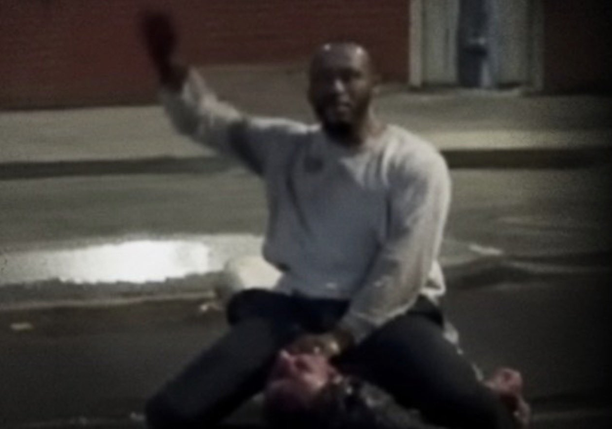 A man sitting on the ground raises his hand, with a blurred background and wet pavement, suggesting a nighttime urban setting.
