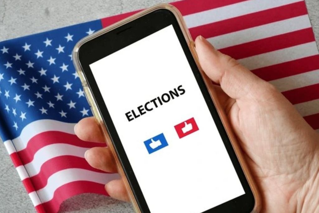 A hand holding a smartphone displaying the word "Elections" with thumbs up icons, set against a backdrop of the American flag.