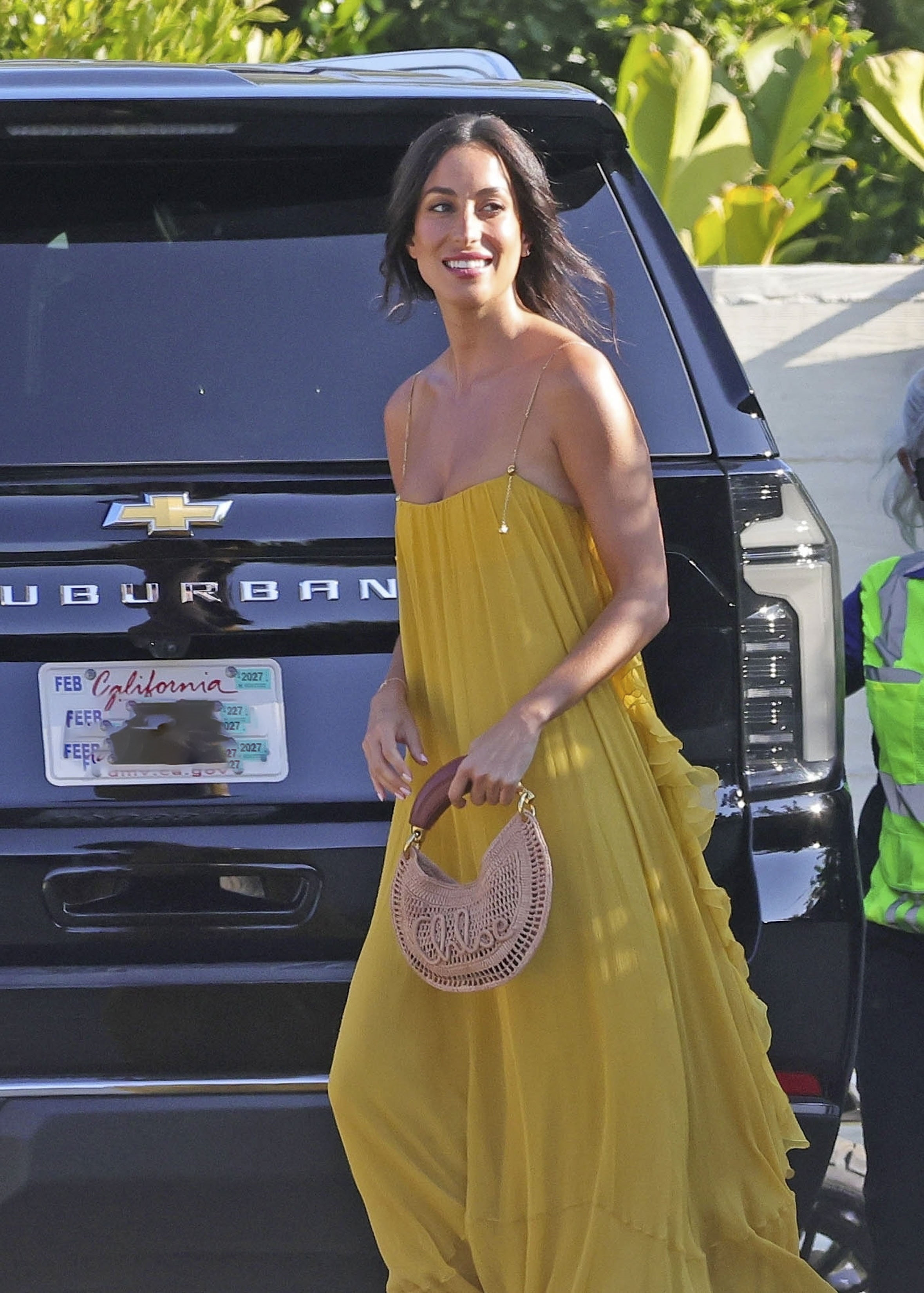 Ines de Ramon smiling in a yellow maxi dress, holding a woven Chloé handbag, walking near a black Chevrolet Suburban in Malibu.