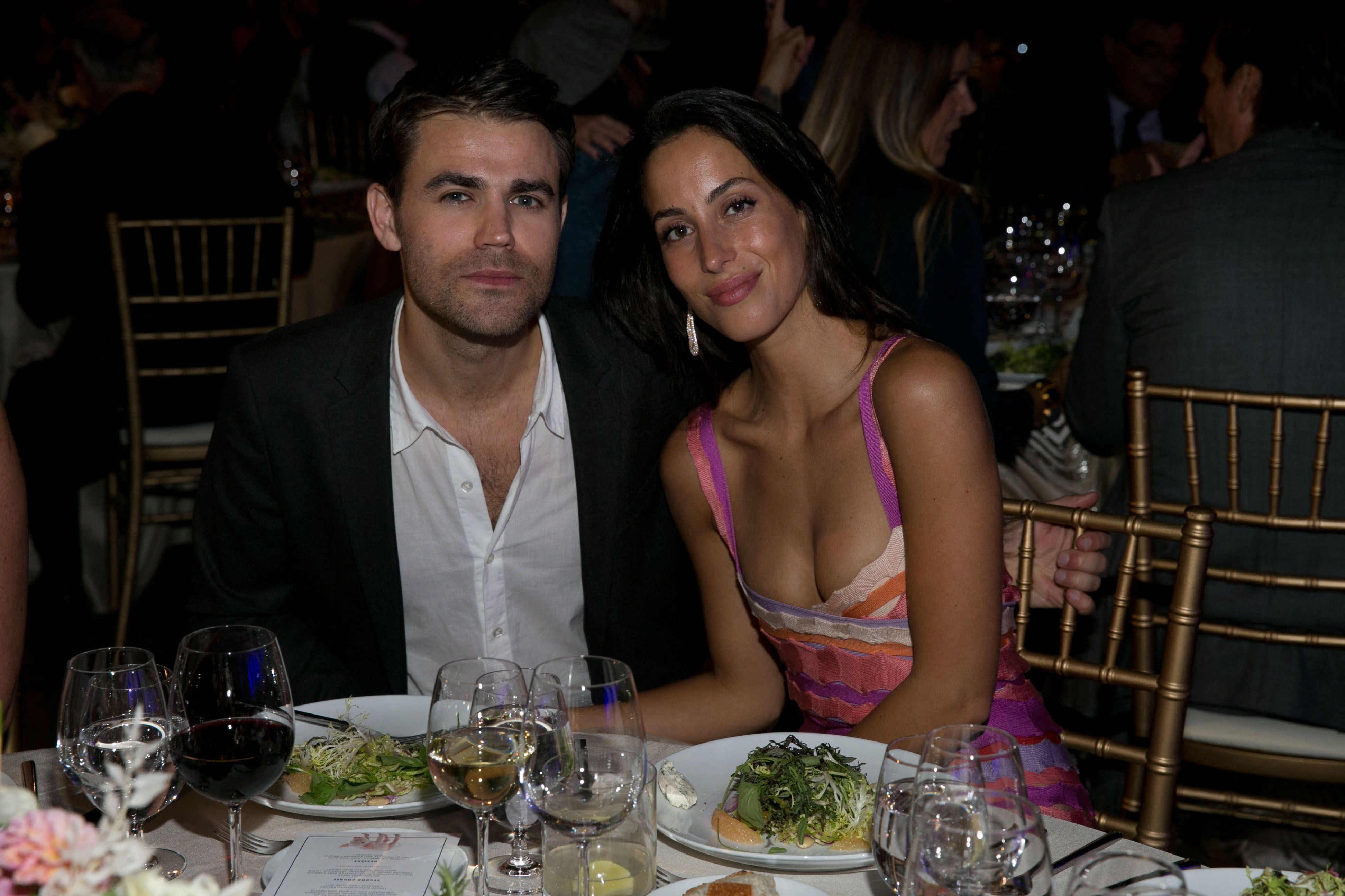 A man in a black blazer and a woman in a colorful striped dress seated together at a formal dinner table with wine glasses and food.