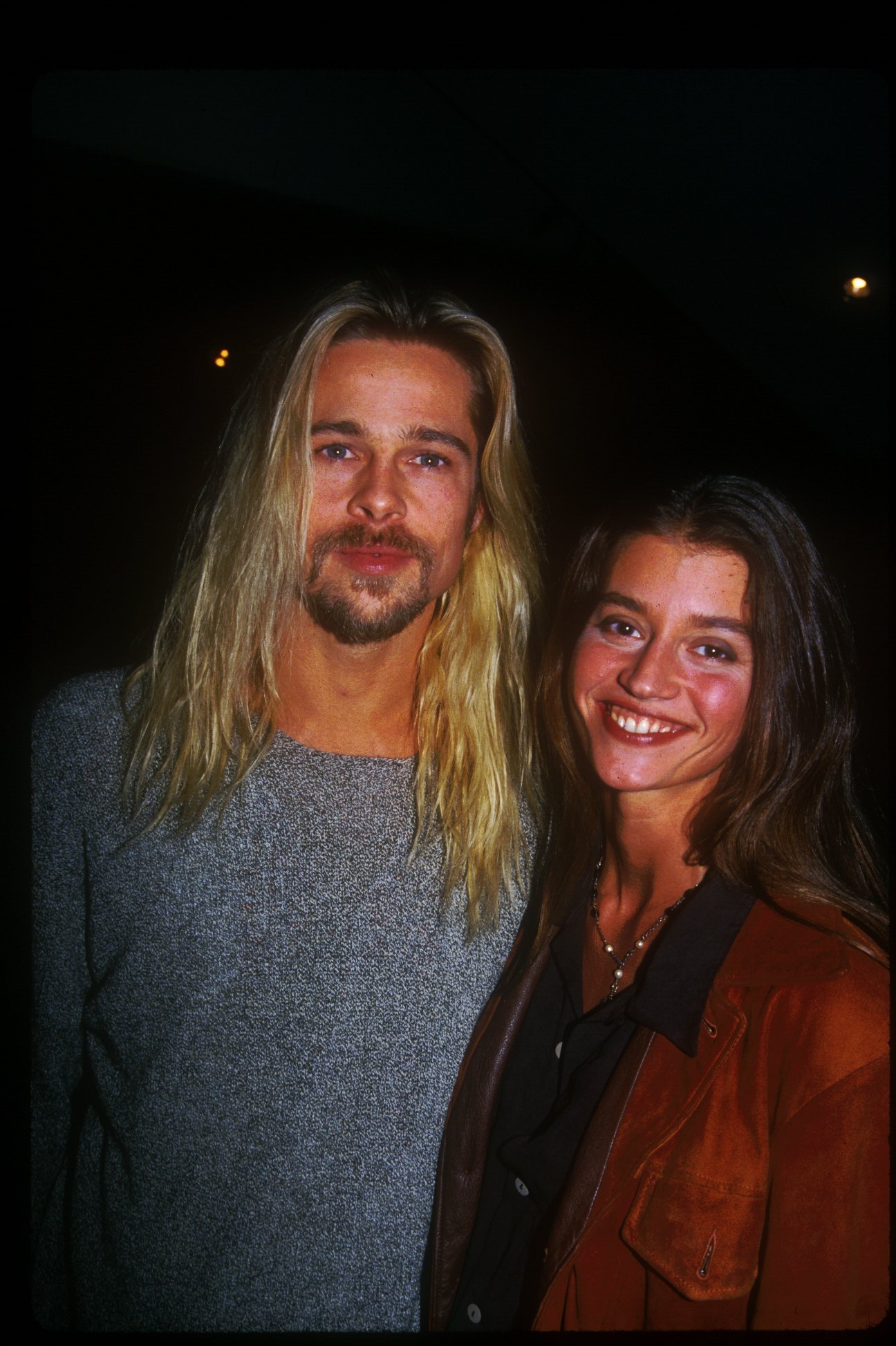 Brad Pitt with long blonde hair and a goatee stands beside a smiling brunette woman in a brown leather jacket at a 1994 Los Angeles event.