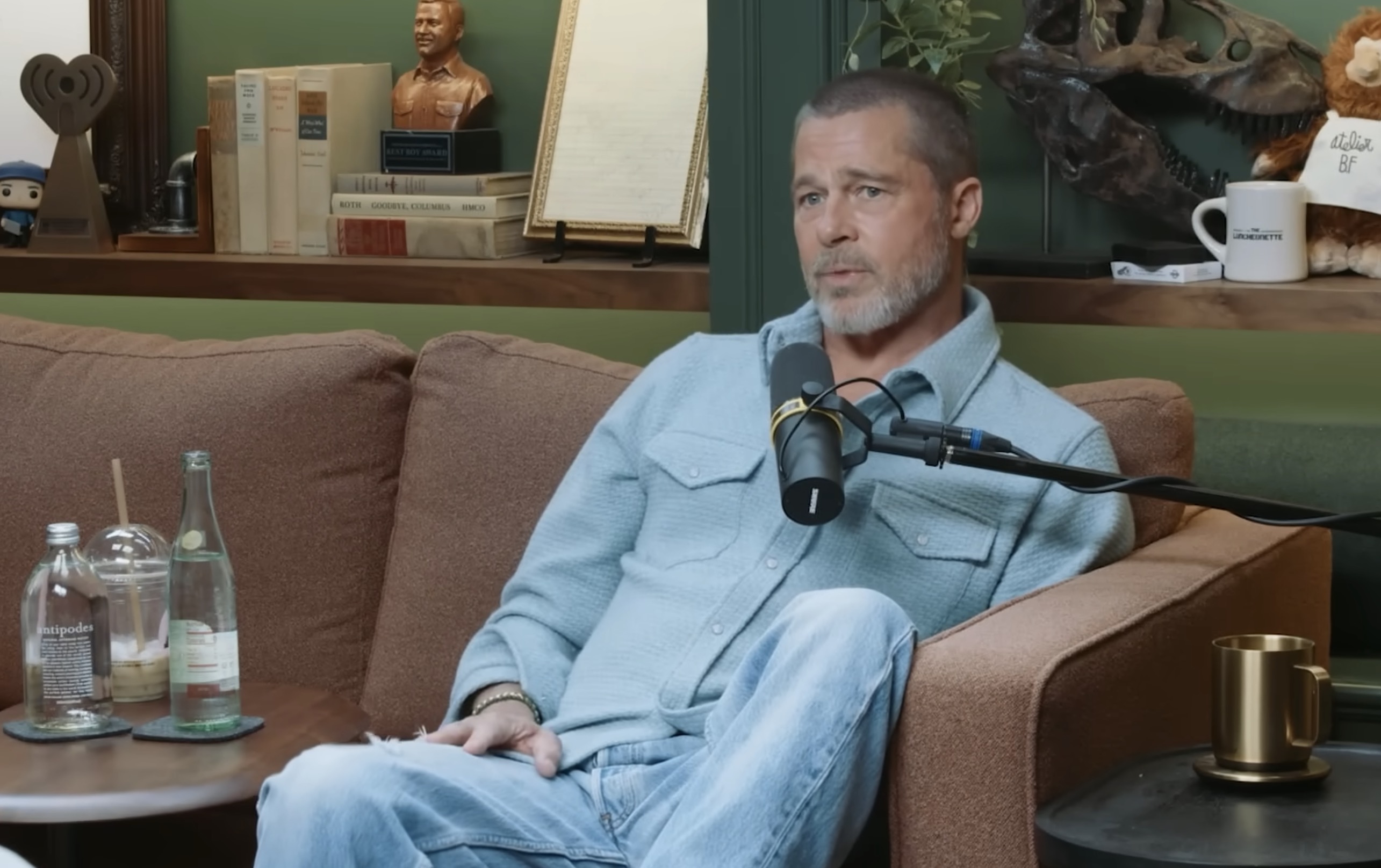 A man with short gray hair and a beard sits on a brown couch wearing a light blue shirt, speaking into a podcast microphone.