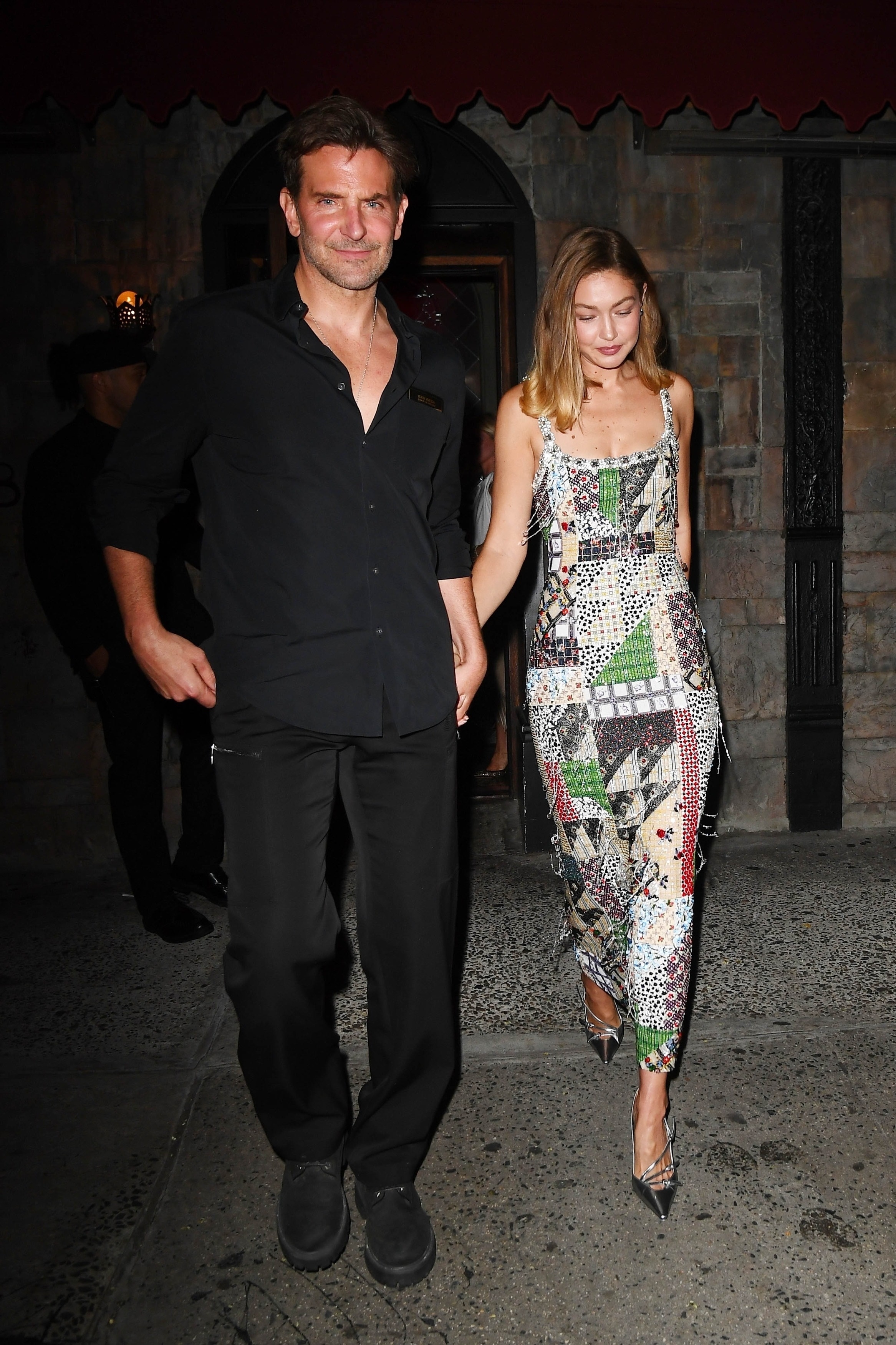 A man in all-black and a woman in a colorful beaded midi dress and silver heels walk hand in hand out of a stone-facade building at night.