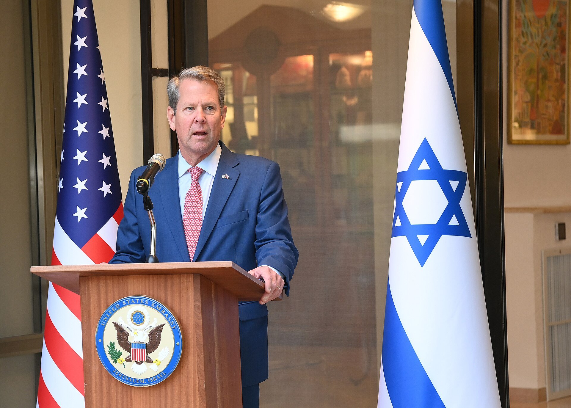 Official speaking event featuring a representative at the United States Embassy in Jerusalem, with American and Israeli flags in the background.