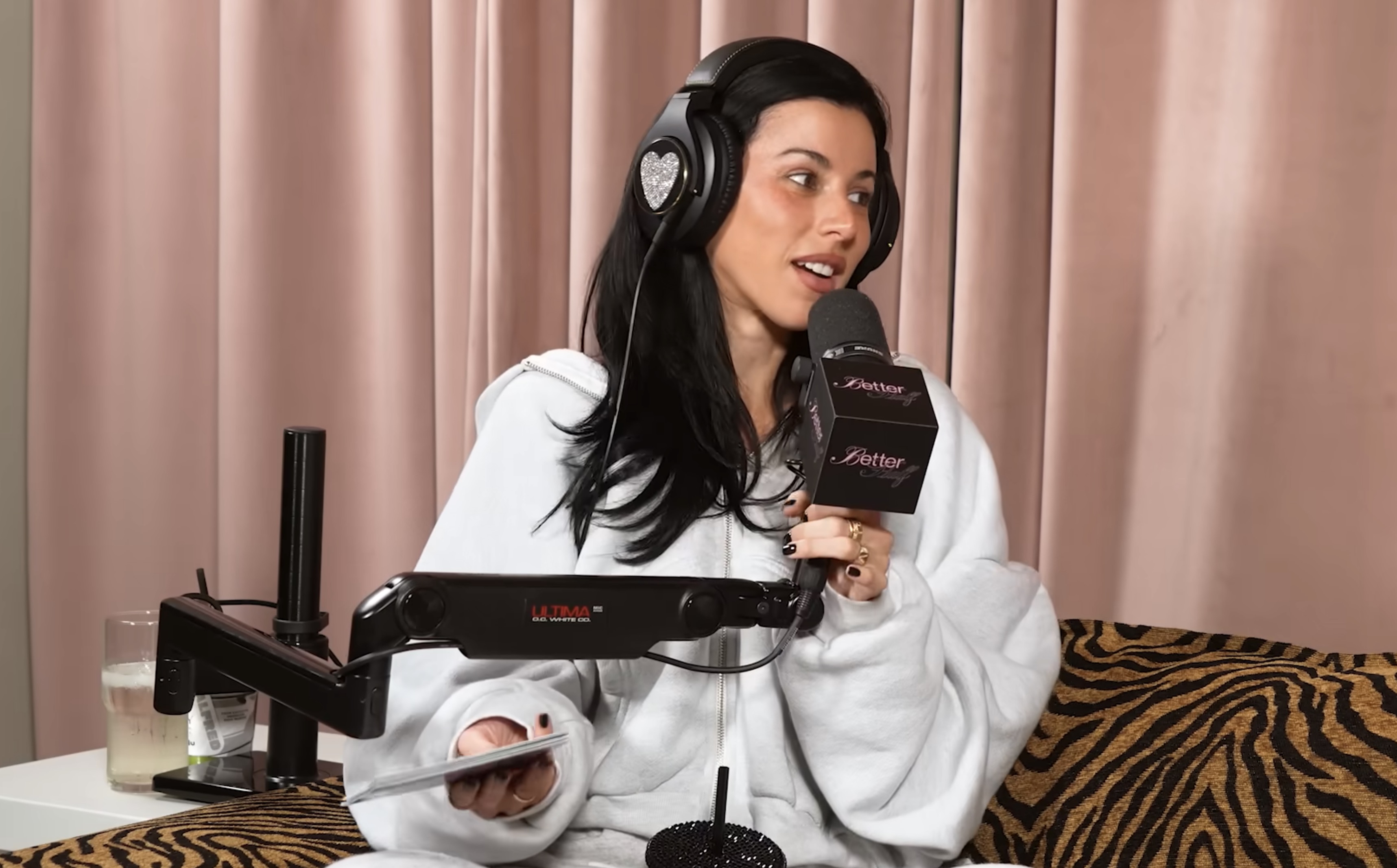 Victoria Villarroel in a white hoodie and bejeweled headphones holding a podcast microphone labeled "Better Half."