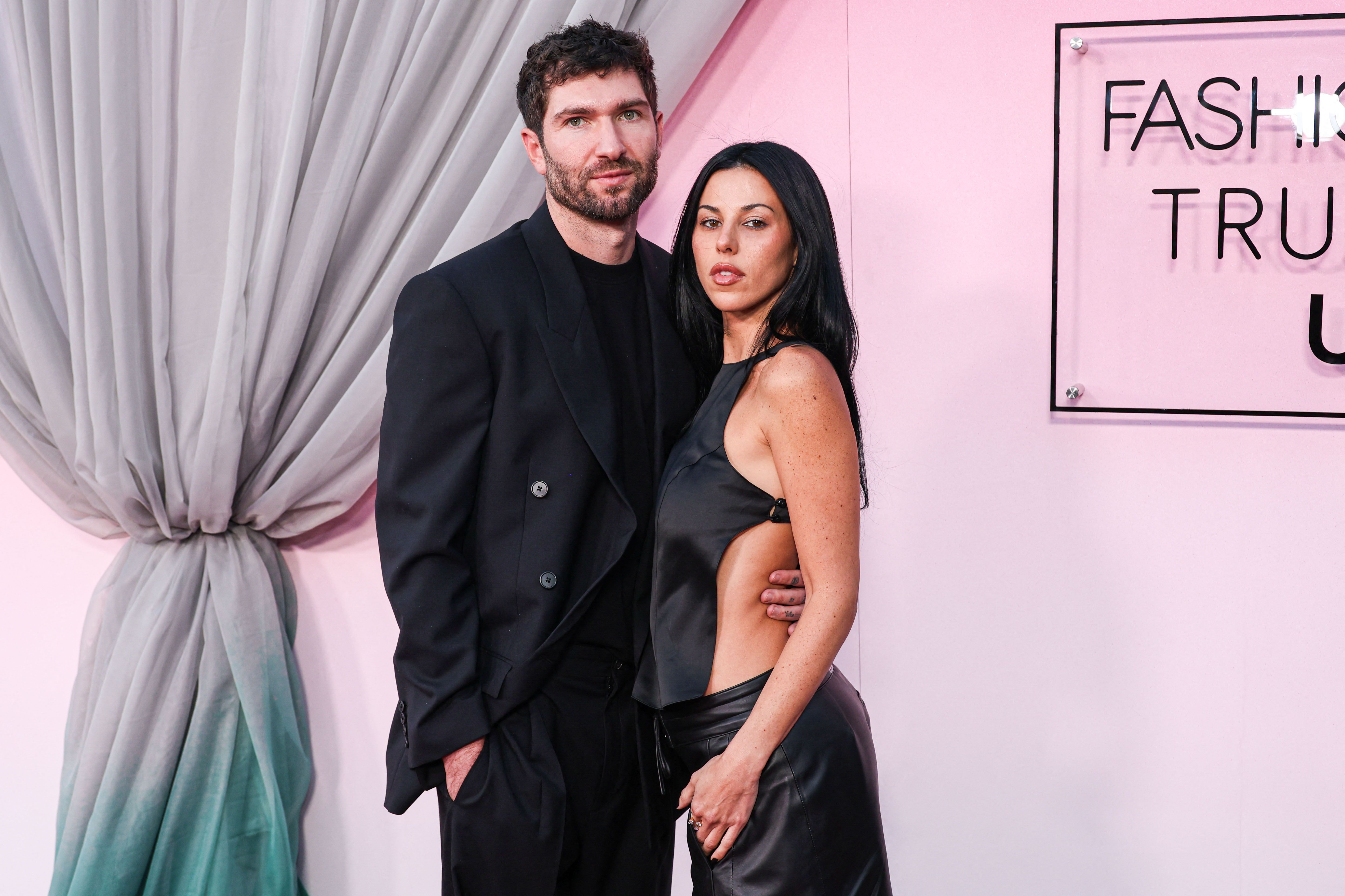 A man in an all-black suit and a dark-haired woman in a black cutout halter top and leather pants posing at a Fashion Trust U.S. Awards event.