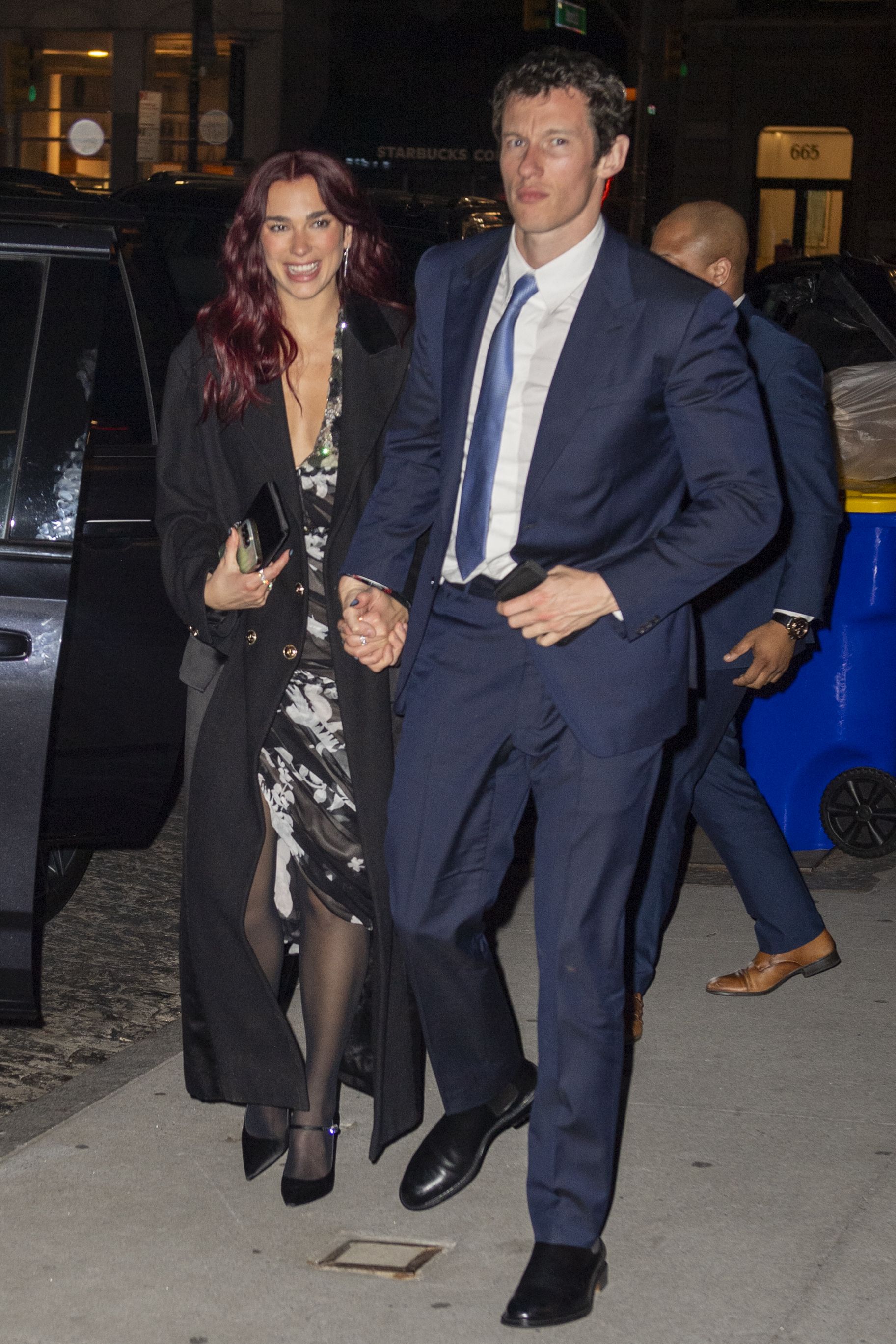 A red-haired woman in a floral dress and black coat walks hand-in-hand with a man in a navy suit on a New York City sidewalk at night.