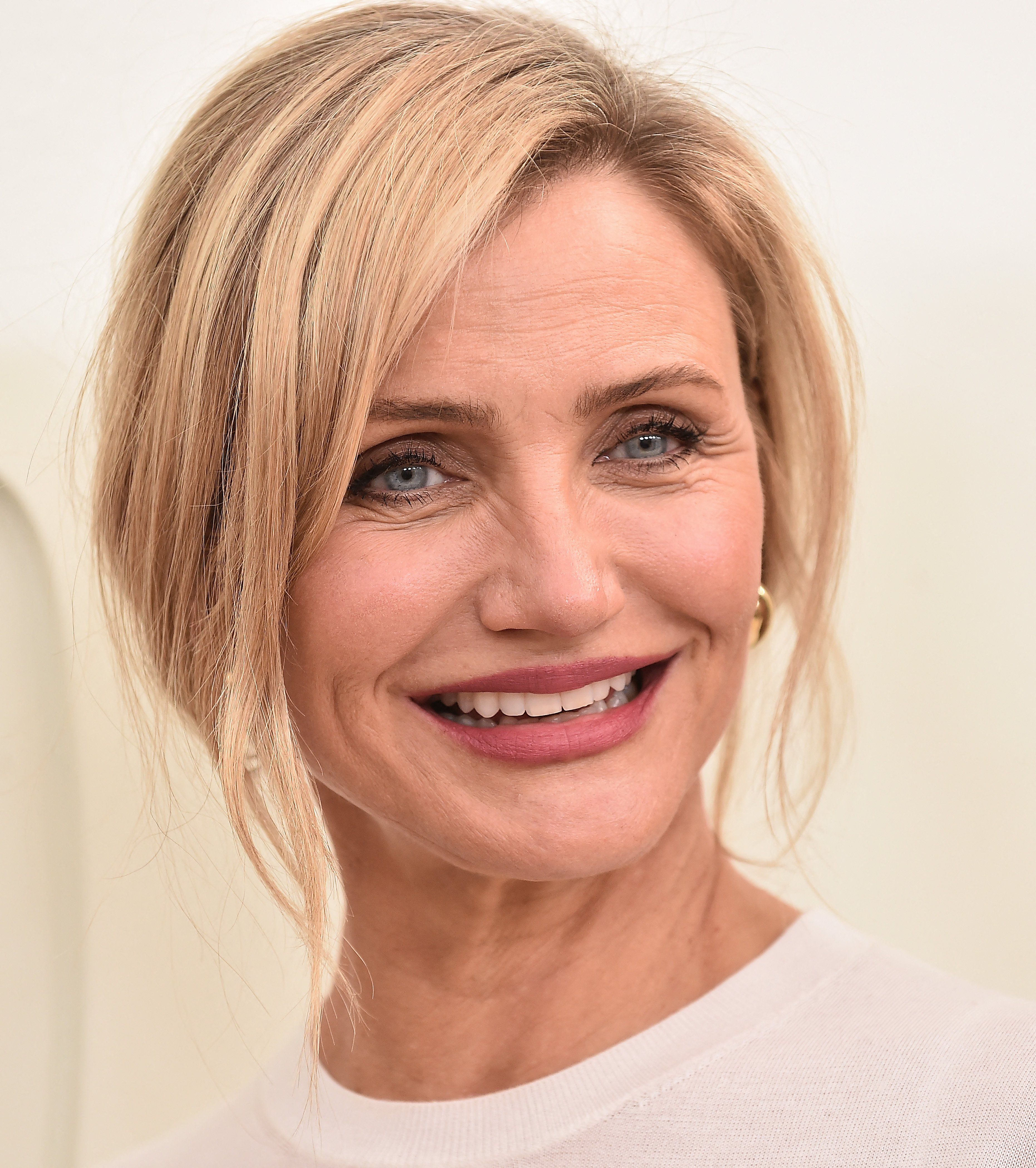 Blonde woman with blue eyes and pink lipstick smiling broadly, hair pulled back loosely, wearing a white top against a light background.