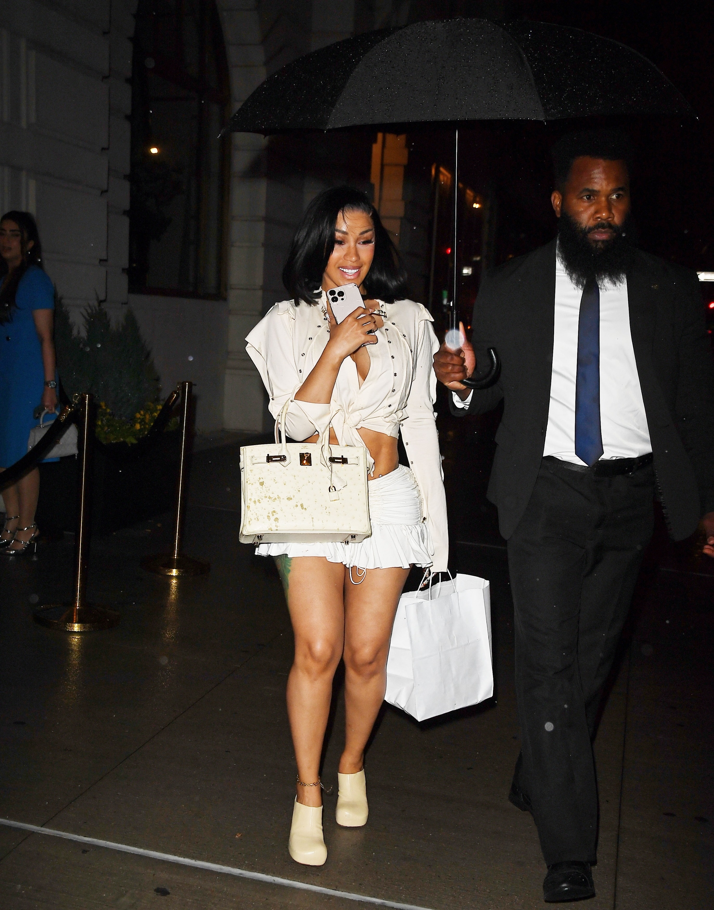 Cardi B in a white knotted top, ruched mini shorts, and cream ankle boots carrying a white Birkin bag outside a NYC restaurant at night under a black umbrella.