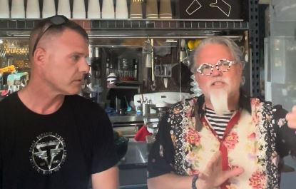Two men engaged in conversation at a bar, one wearing a floral shirt and glasses, discussing cocktails and mixology techniques.