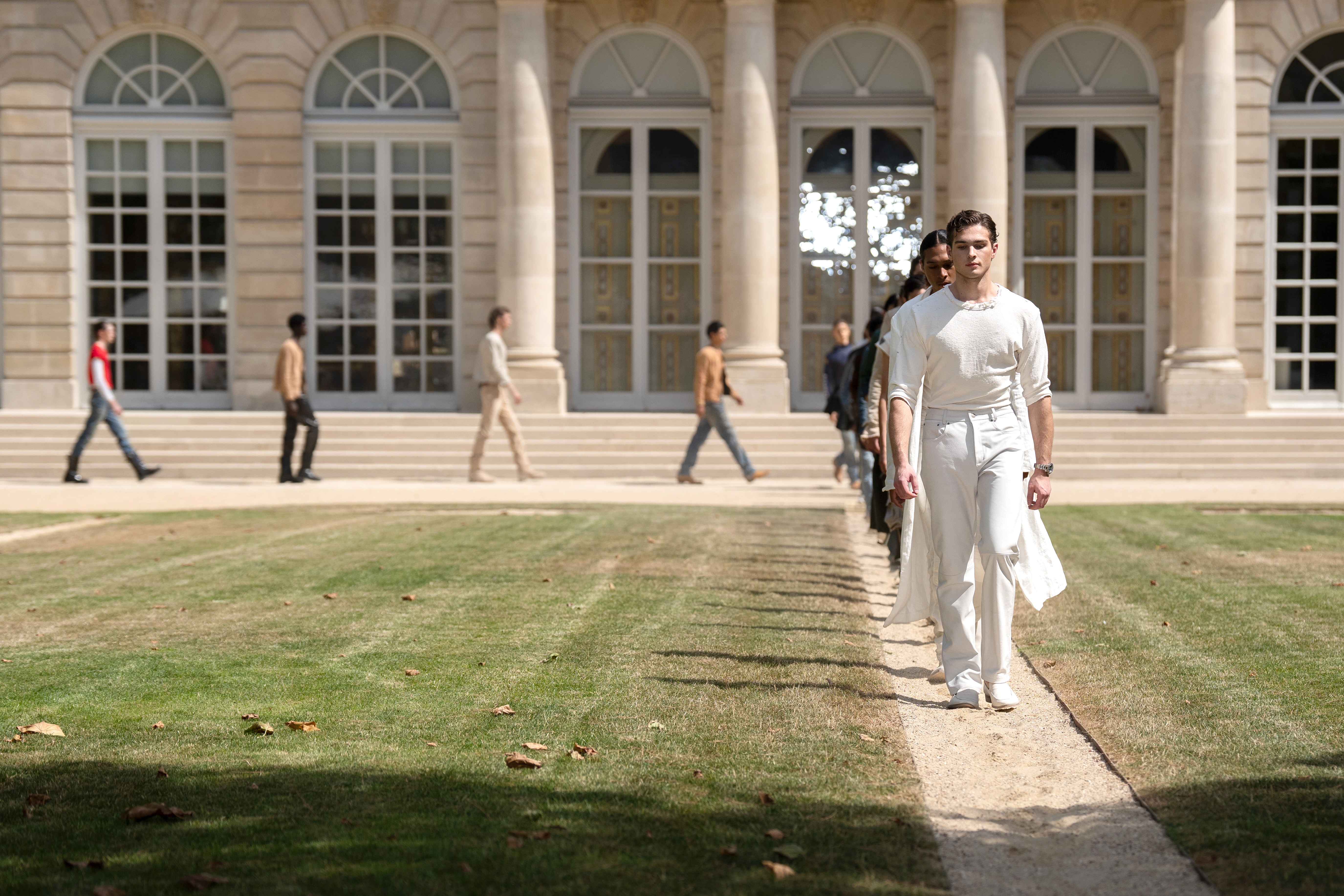 A male model in an all-white outfit leads a line of models walking along a path in front of a neoclassical building during an outdoor fashion show.