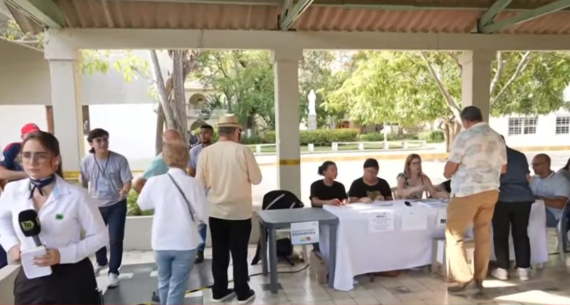 Voters engage with officials at an outdoor polling station, with tables set up for registration and assistance in a community setting.
