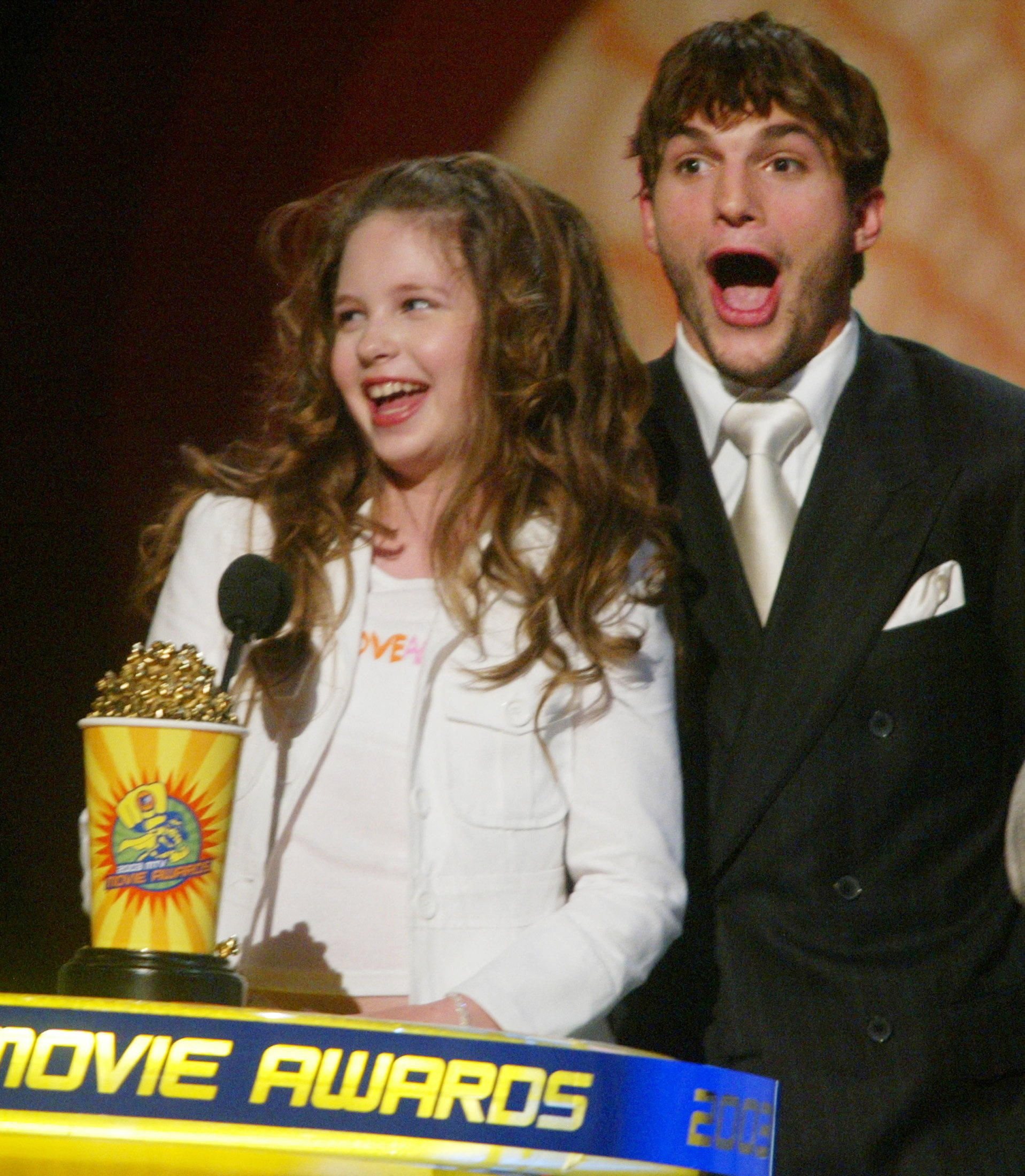 A young curly-haired girl and a man in a black suit with a white tie stand at the MTV Movie Awards podium, both with mouths open in excitement.