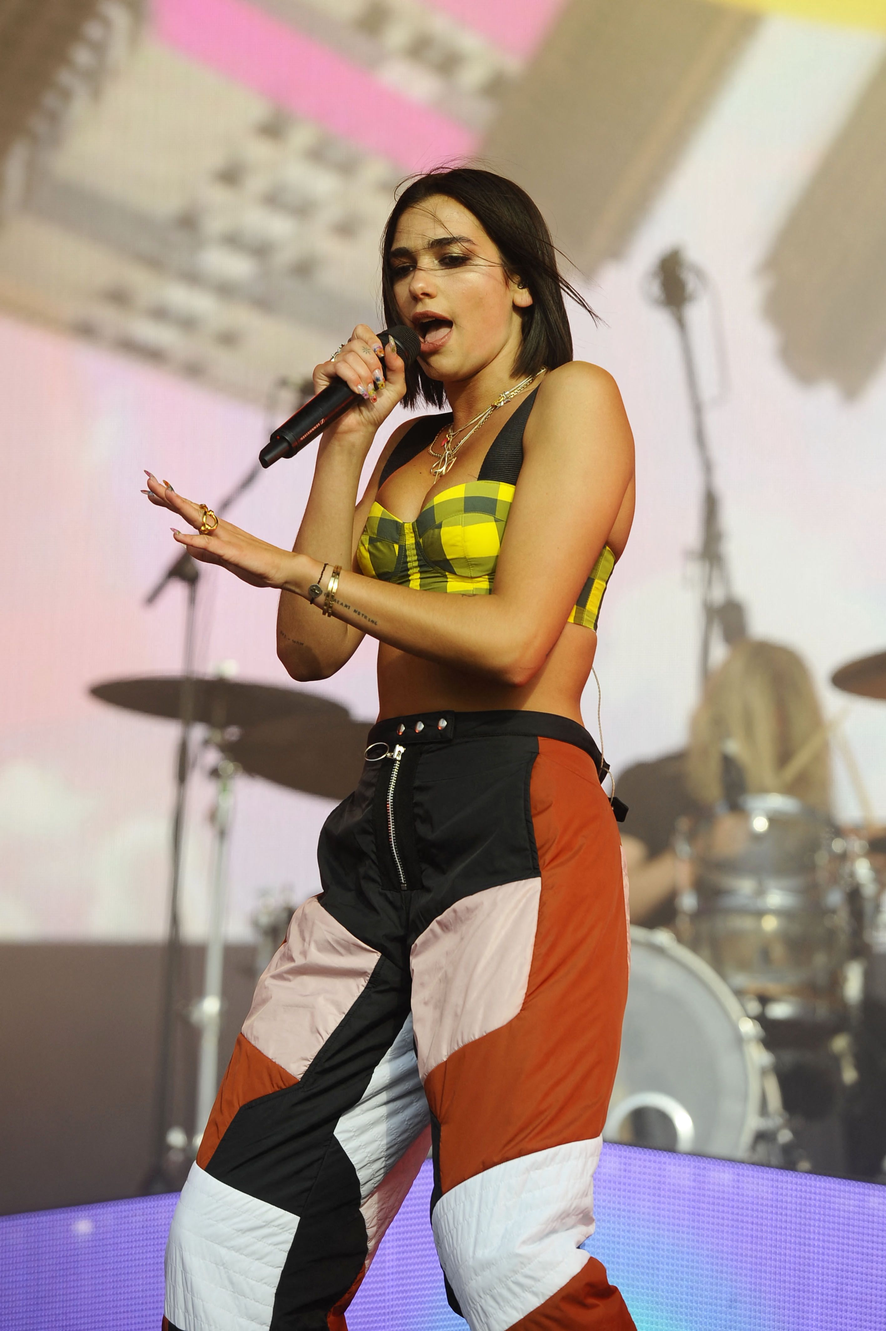 Female singer performing on stage, holding a microphone, wearing a yellow plaid crop top and colorblock pants, with a drum kit visible in the background.