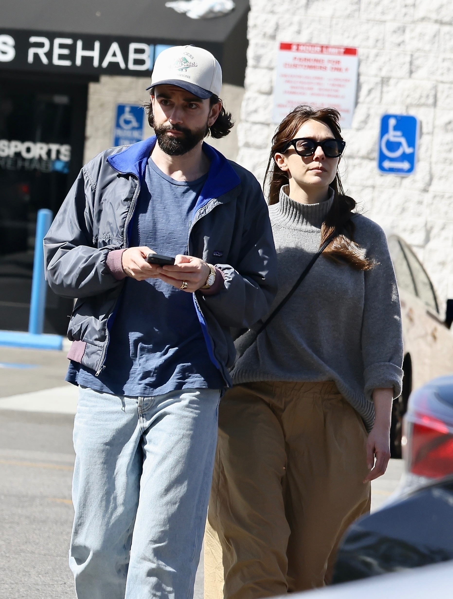 A bearded man in a navy jacket, baseball cap, and jeans walks alongside a woman in a gray turtleneck sweater, tan pants, and dark sunglasses outside a sports rehab building.