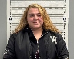 Mugshot of a woman with curly blonde hair wearing a black jacket, standing in front of a height chart.