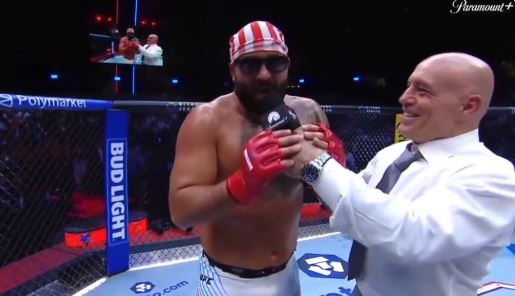 Fighter in red and white striped headband and sunglasses celebrates victory in the octagon while being interviewed by a commentator.
