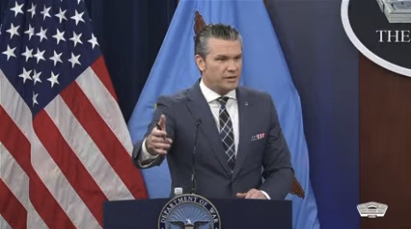 Official delivering a statement at a press conference in front of the U.S. flag and Department of Defense backdrop.