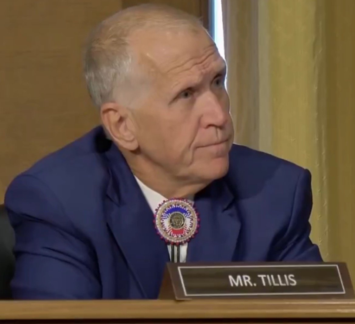 Senator Tillis attentively listening during a committee meeting, wearing a blue suit and a decorative pin.