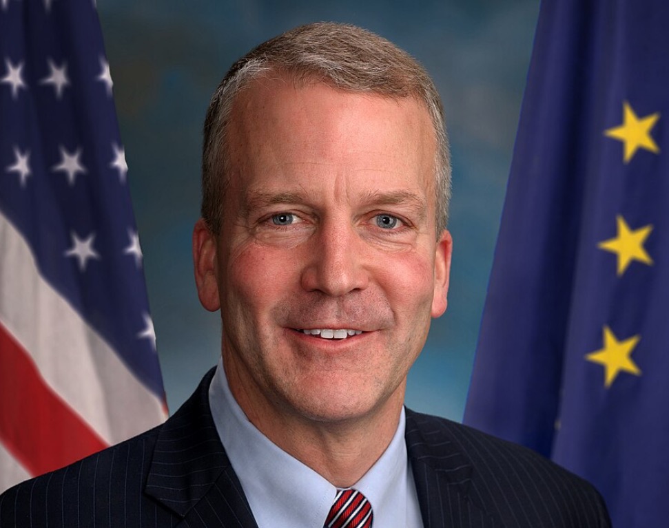 Portrait of a smiling man in a suit, standing in front of the American and European Union flags.