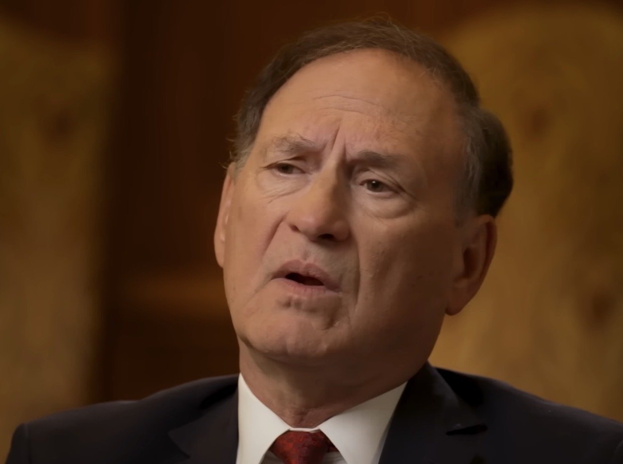 Man in a suit speaking thoughtfully, with a blurred background, conveying a serious discussion or interview setting.