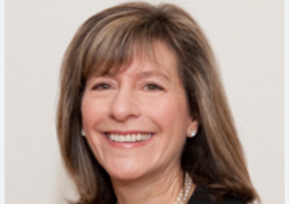 Smiling woman with shoulder-length brown hair, wearing earrings and a necklace, against a neutral background.